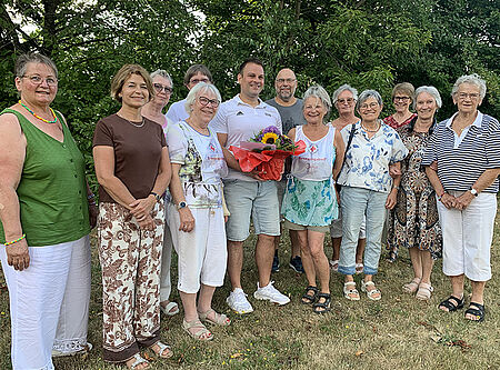 Gruppe von älteren und einem jüngeren Menschen im Freien, der jüngere Mann hält einen Blumenstrauß