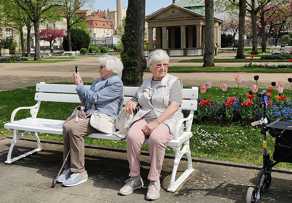 Zwei ältere Frauen sitzen auf einer weißen Parkbank vor einem Blumenbeet mit Tulpen und einem Pavillon im Hintergrund.