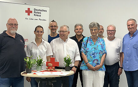 Gruppe von acht Personen steht hinter einem runden Tisch mit Pflanzen, einem Stoffbären und Papieren vor einem Banner des Deutschen Roten Kreuzes