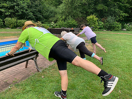 Drei Personen stützen sich auf einer Wiese mit ausgestrecktem Bein an einer Holzbank ab
