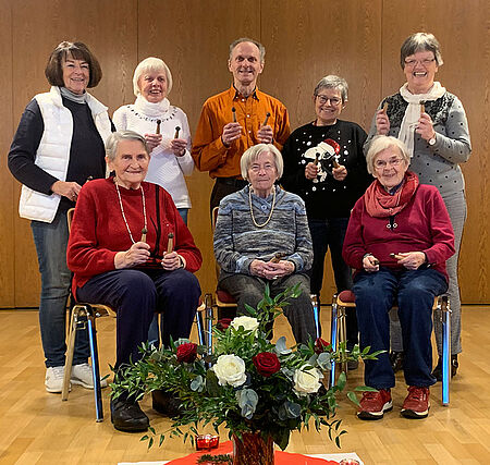 Gruppe von sieben Personen, drei sitzen auf Stühlen, vier stehen dahinter, alle halten kleine Glöckchen, davor ein Blumenarrangement auf dem Boden.